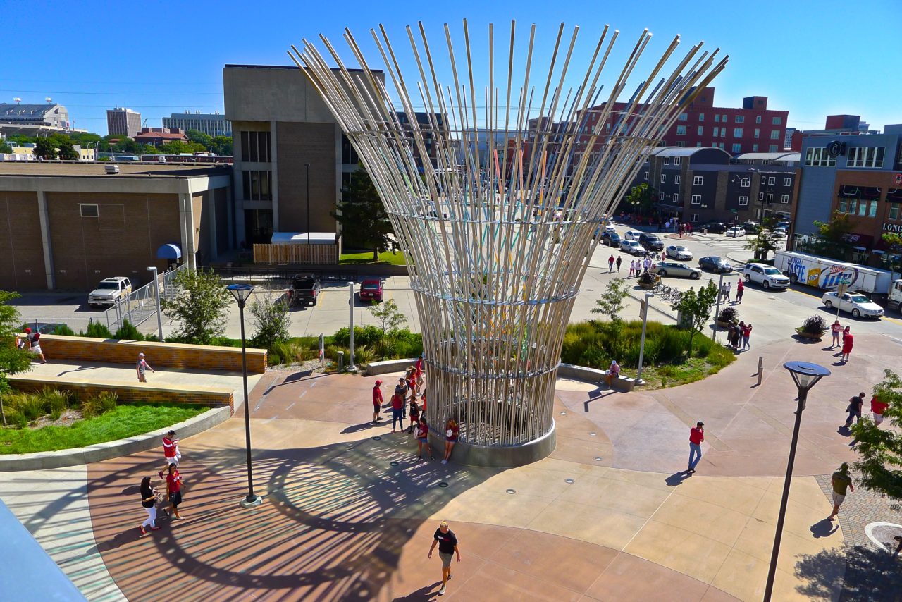 Harvest monumental public sculpture in Lincoln, Nebraska day view, showing dichroic glass patterns on the plaza sidewalk amid fans waiting for the upcoming football game. | Image 2 | Ed Carpenter, Artist