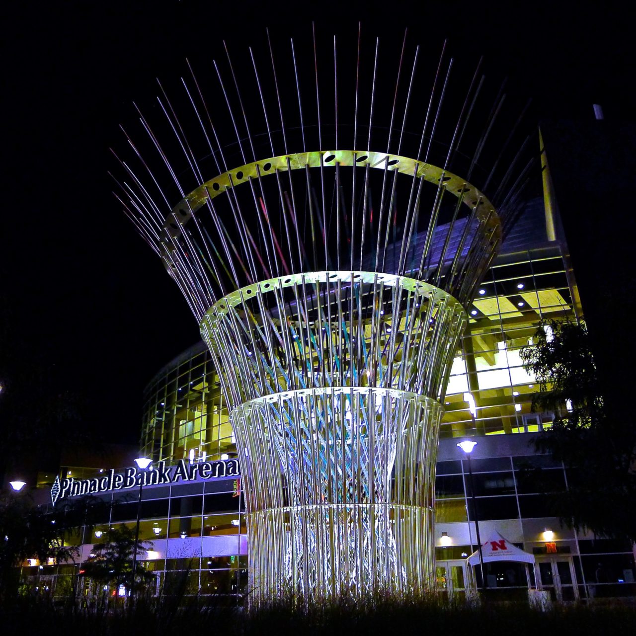 Harvest monumental public sculpture in Lincoln, Nebraska with night lighting in the plaza in front of the Pinnacle Bank Arena amplifies the stainless steel and dichroic glass structural tension elements crisscrossing in the center of the sculpture. | Image 1 | Ed Carpenter, Artist